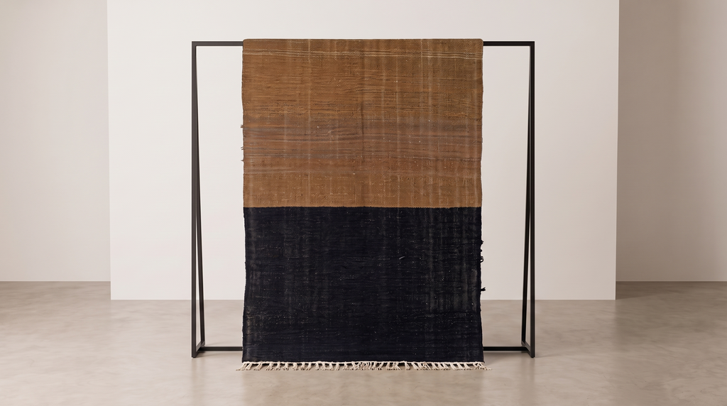 A woven textile throw blanket with a horizontal color-block design, featuring a warm tan top half and a dark charcoal-black bottom half, hanging from a minimalist black metal frame in a clean studio setting.