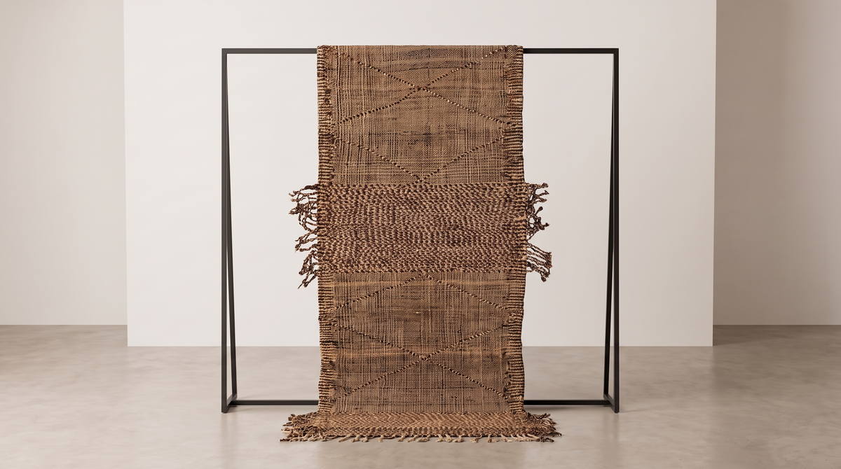 A brown woven textile throw blanket with fringe details, draped over a minimalist black metal frame in a clean, modern studio setting.