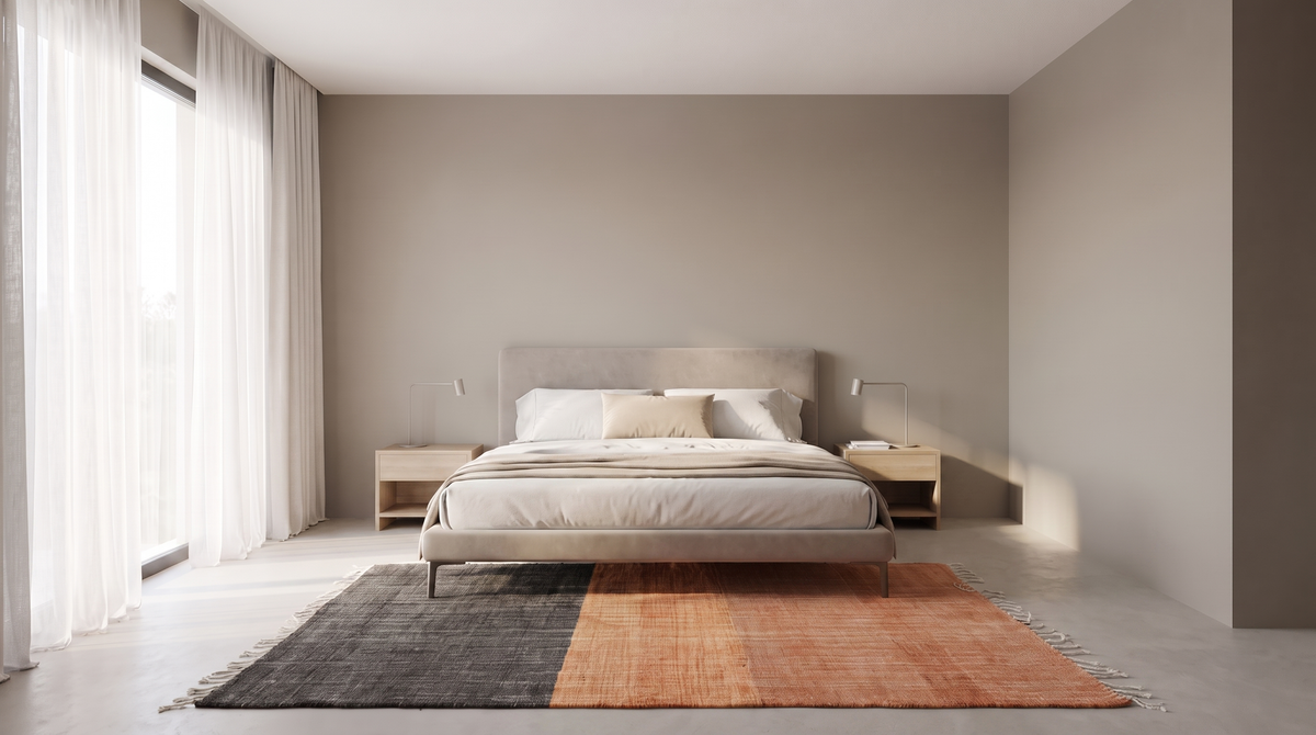 A modern minimalist bedroom featuring a low-profile bed with neutral beige bedding, set against a warm taupe-gray wall. The bed is styled with two light wood nightstands, a matching headboard, and a large rectangular area rug in black and terracotta orange tones.
