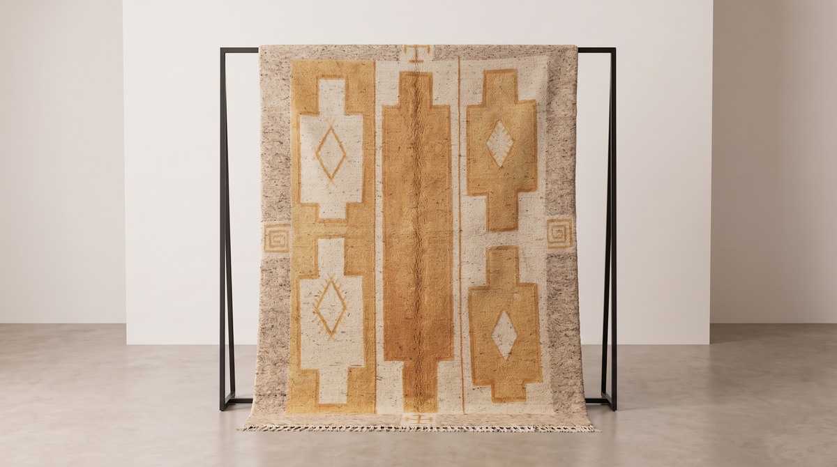 A woven area rug with a Southwestern geometric pattern in beige, tan, and cream, draped over a minimalist black metal stand in a clean, modern studio setting.