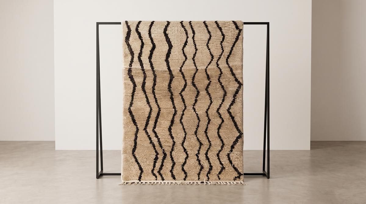 A woven throw blanket with a beige base and black zigzag pattern, draped over a minimalist black metal stand in a clean studio setting.