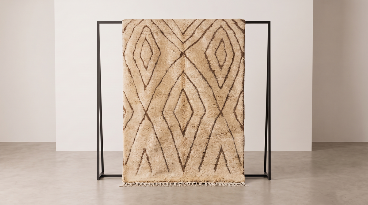 A beige shag area rug with a dark brown geometric diamond pattern, draped over a minimalist black metal stand in a clean studio setting.