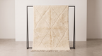 A cream-colored textured throw blanket with a diamond quilted pattern and fringe trim, draped over a minimalist black metal stand in a clean, minimalist studio setting.