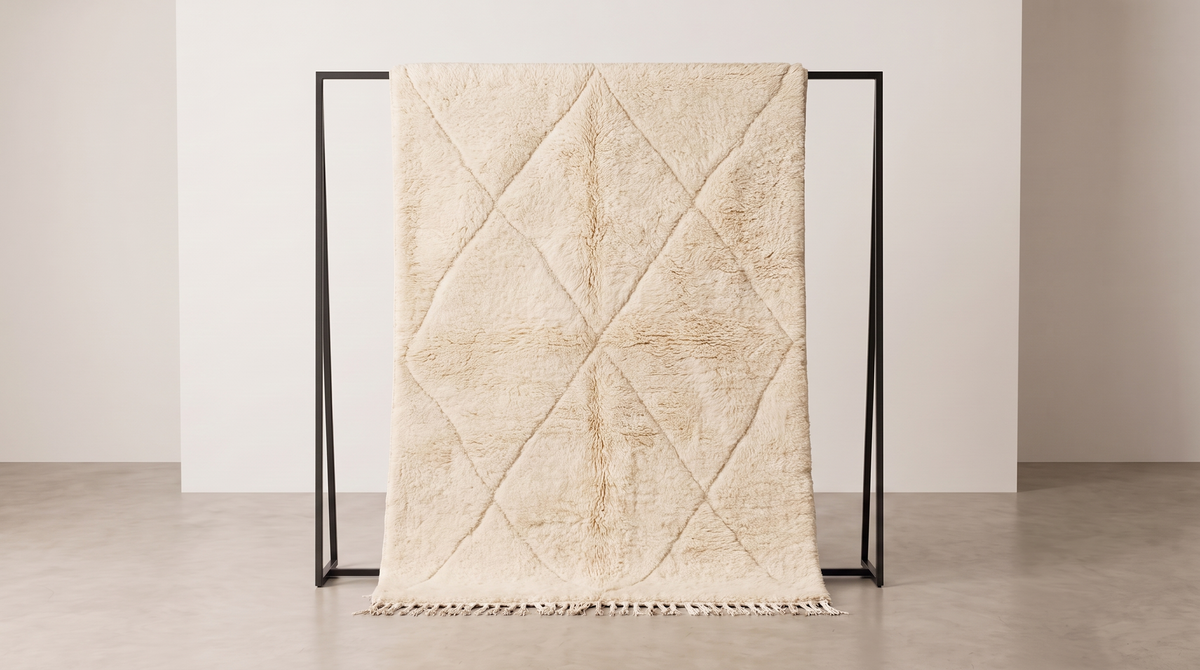 A cream-colored textured throw blanket with a diamond quilted pattern and fringe trim, draped over a minimalist black metal stand in a clean, minimalist studio setting.