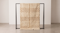 A woven throw blanket with a beige and brown geometric pattern is draped over a minimalist black metal stand in a clean, modern room setting.
