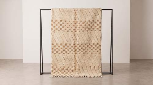 A woven throw blanket with a beige and brown geometric pattern is draped over a minimalist black metal stand in a clean, modern room setting.