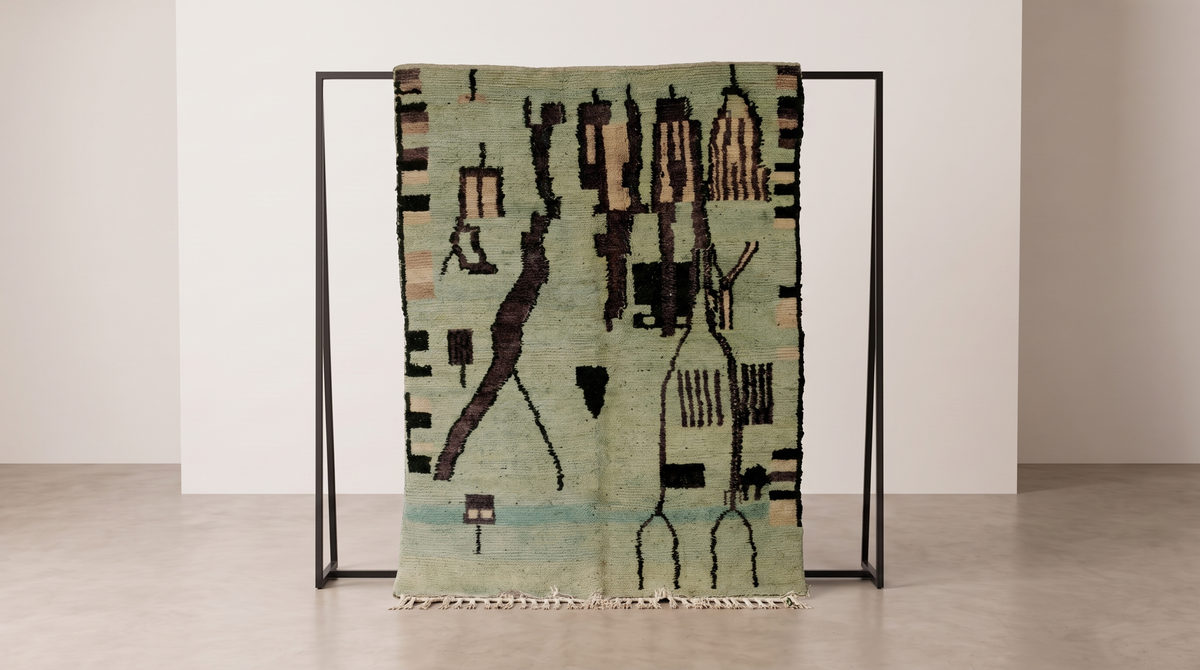 Boujad Bloom rug detail with abstract tribal figures in muted green, beige, black, and light blue, displayed on a stand in a studio.