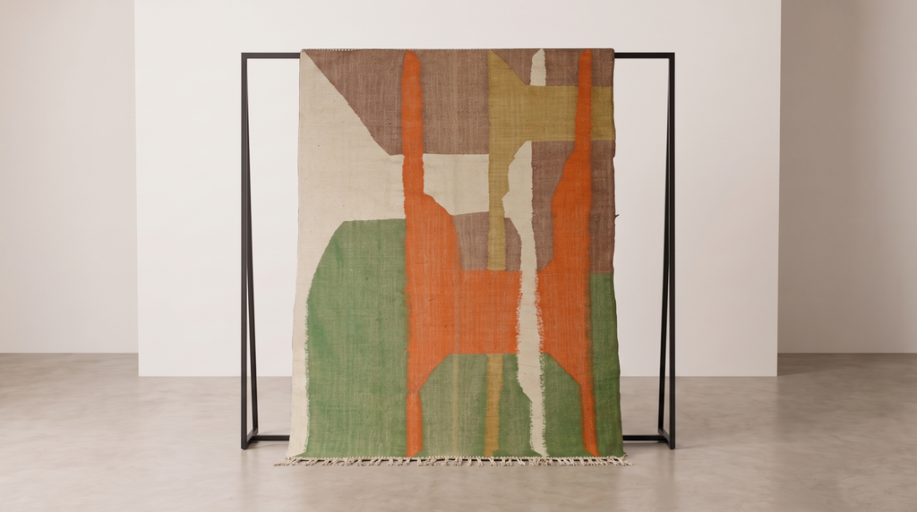 An abstract area rug with geometric shapes in orange, green, brown, and cream, draped over a minimalist black metal stand in a clean, modern room.